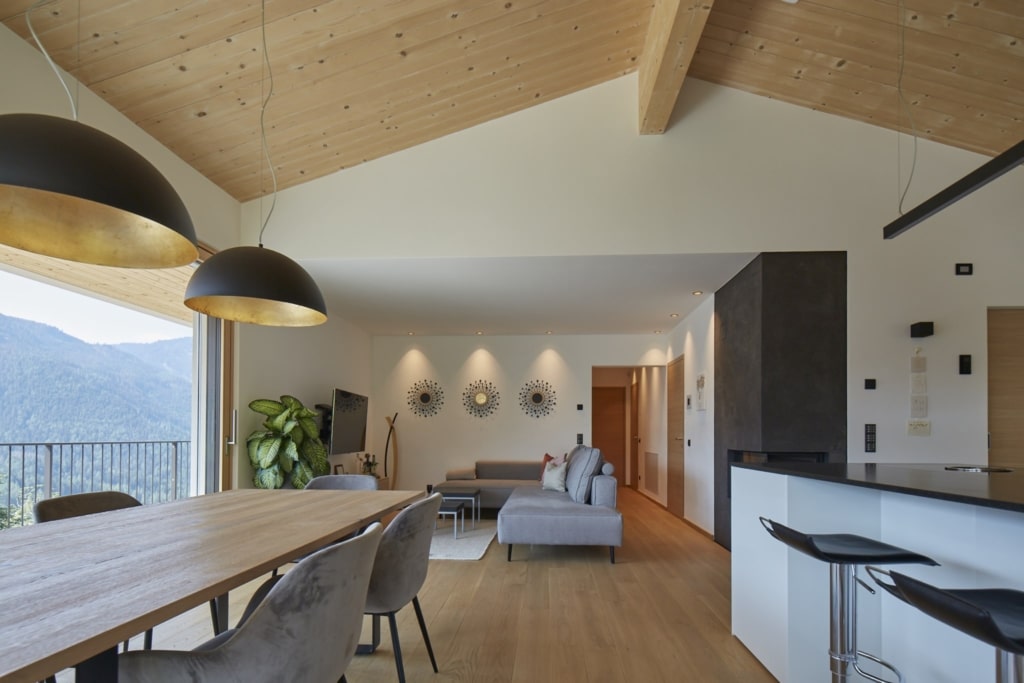 Una zona giorno luminosa e aperta con un soffitto in legno massello Holzius, rivestito con pannelli di legno chiaro e attraversato da una trave di legno. Il pavimento è rivestito con assi di legno. Due grandi lampade a sospensione nere con l'interno dorato illuminano un tavolo di legno con sedie grigie. Attraverso un'ampia vetrata lo sguardo spazia su un paesaggio verde con le montagne. Una grande pianta da appartamento è posizionata accanto alla vetrata. Sullo sfondo si trova un salotto con un divano grigio e un televisore. Un camino nero si trova tra la zona giorno e la zona pranzo. Lo stile Holzius crea un'atmosfera calda e accogliente. La combinazione di legno ed elementi moderni conferisce alla stanza un tocco elegante e accogliente.