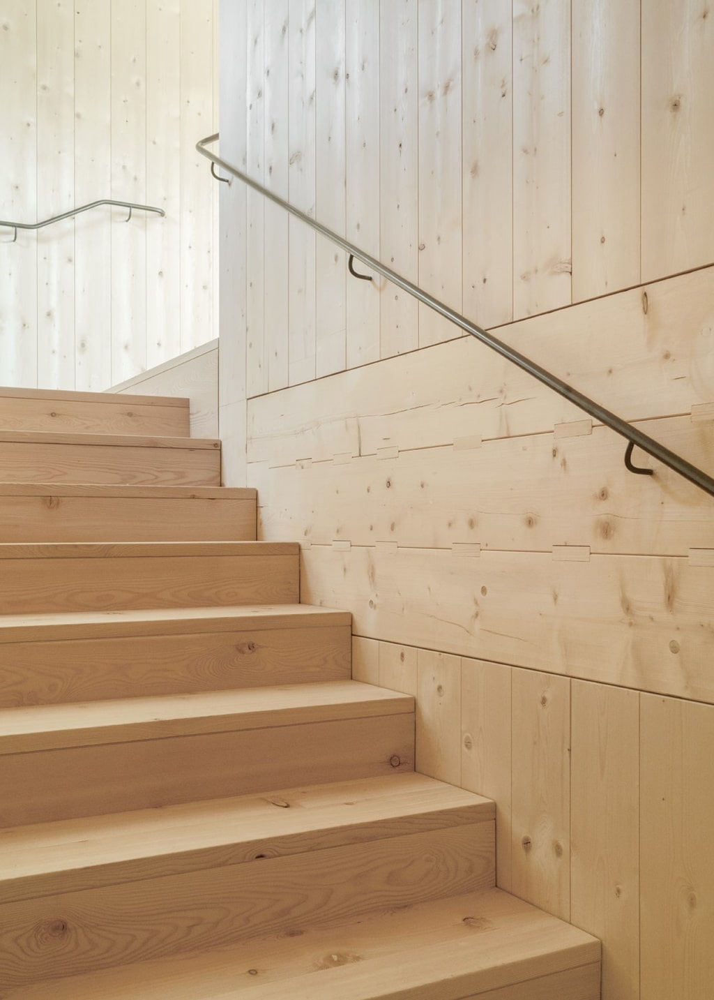 Una zona scale dal design pulito, realizzata in legno, che trasmette un'immagine calda e serena: gradini in legno massello e una parete composta da elementi Holzius con giunti a vista caratterizzano l'ambiente. Una semplice ringhiera in metallo accompagna la scala e sottolinea lo stile essenziale e artigianale dell'opera.