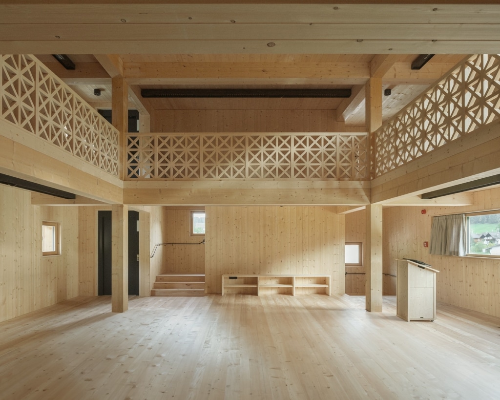 Un ampio spazio interno a due piani, interamente realizzato in legno, che trasmette una sensazione di chiarezza e serenità: pilastri e travi massicci scandiscono lo spazio aperto, mentre una galleria perimetrale con una ringhiera in legno dai motivi geometrici e delicati delimita il livello superiore. Una piccola scala, sedute integrate e un bancone dal design sobrio strutturano lo spazio in modo funzionale, mentre ampie aperture e finestre lasciano entrare la luce naturale.