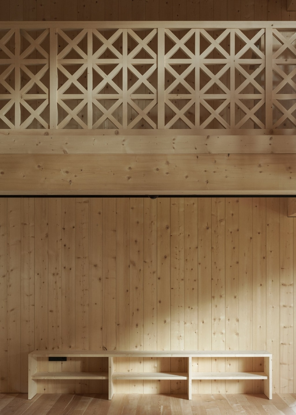 Un interno interamente realizzato in legno, caratterizzato da un design pulito e sobrio: un'alta parete rivestita verticalmente è interrotta da una galleria dotata di una ringhiera dal design delicato e dai motivi geometrici. Sotto di essa, una semplice panca integrata corre lungo la parete, mentre la luce naturale mette in risalto la calda superficie del legno.