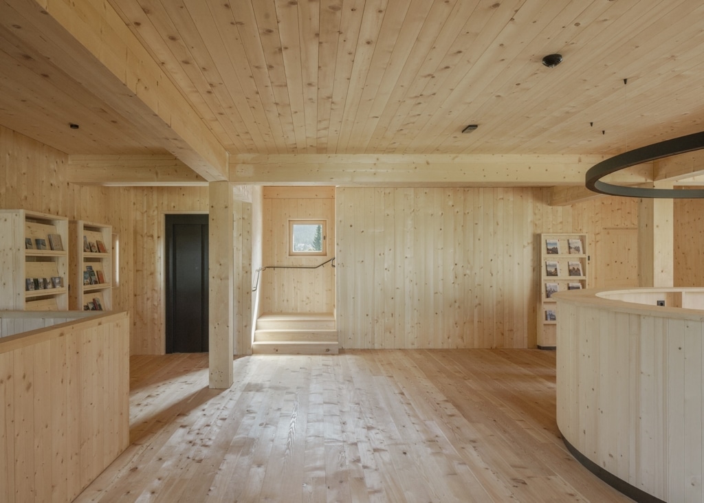 Un interno luminoso e aperto, interamente realizzato in legno, che crea un'atmosfera sobria e tranquilla: pareti, soffitto e pavimento sono interamente rivestiti in legno chiaro e scanditi da pilastri e travi massicci. Un bancone circolare rivestito in legno, con una lampada ad anello sospesa sopra di esso e scaffali integrati, definisce lo spazio, mentre una piccola scala e aperture strategicamente posizionate collegano le diverse zone.