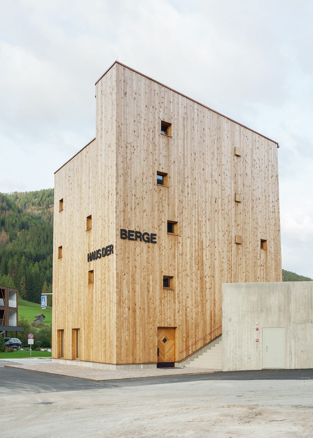 Un edificio compatto in legno, caratterizzato da un volume chiaro e monolitico e da una facciata articolata verticalmente, domina lo spazio stradale: le aperture delle finestre disposte in modo irregolare e la scritta «HAUS DER BERGE» definiscono la superficie. Un edificio in cemento armato adiacente sul lato, dotato di una scala esterna, e l’area d’ingresso al piano terra completano il linguaggio architettonico sobrio e preciso.