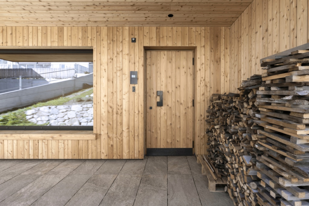 Una vista esterna di una moderna casa Holzius con una facciata composta da doghe di legno verticali e un tetto spiovente. L'edificio presenta ampie finestre che lasciano entrare molta luce naturale e un balcone coperto con ringhiera in legno. In primo piano si trova un giardino ben curato con fiori colorati e un piccolo prato. Il cielo รจ blu con poche nuvole, il che crea un'atmosfera accogliente e invitante.