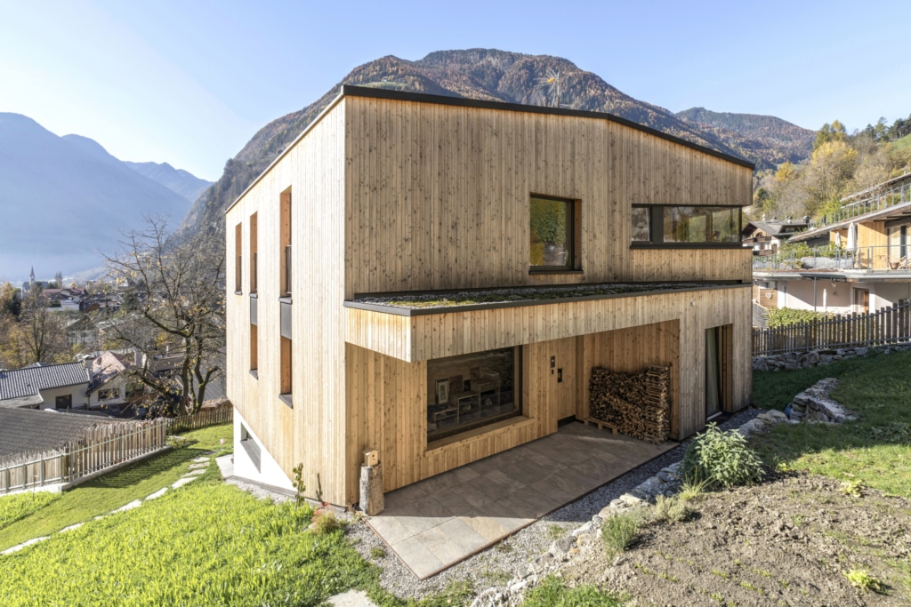 Una moderna casa in legno con una facciata composta da doghe verticali, situata in un pittoresco paesaggio montano. L'edificio presenta ampie finestre che lasciano entrare molta luce naturale e un tetto spiovente. In primo piano si vede una zona pavimentata che conduce alla porta d'ingresso. Il giardino รจ verde e ben curato, con una piccola catasta di legna sul lato. Sullo sfondo si ergono dolci colline e montagne sotto un cielo azzurro e limpido, creando un'atmosfera tranquilla e accogliente.