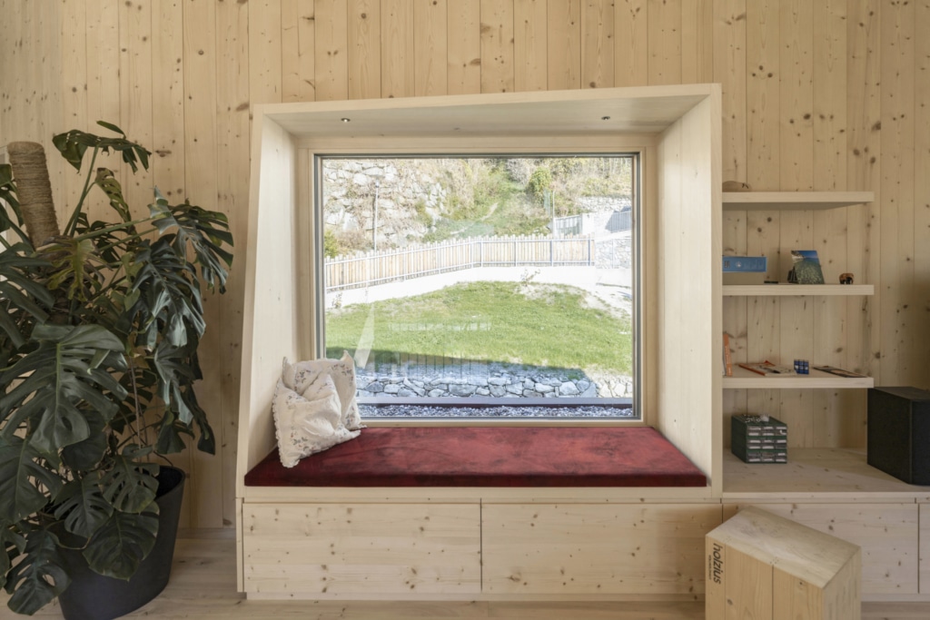 Una finestra con seduta in un ambiente moderno con pareti in legno massello Holzius che mettono in risalto le venature naturali del legno. La finestra รจ ampia e offre una vista su un prato verde. La seduta รจ dotata di un cuscino rosso e dispone di uno spazio di stivaggio sottostante. Sul lato sinistro si trova una grande pianta in un vaso nero, mentre sul lato destro รจ visibile una mensola con vari oggetti decorativi. L'atmosfera complessiva รจ luminosa e accogliente, esaltata dalla luce del giorno che filtra dalla finestra.