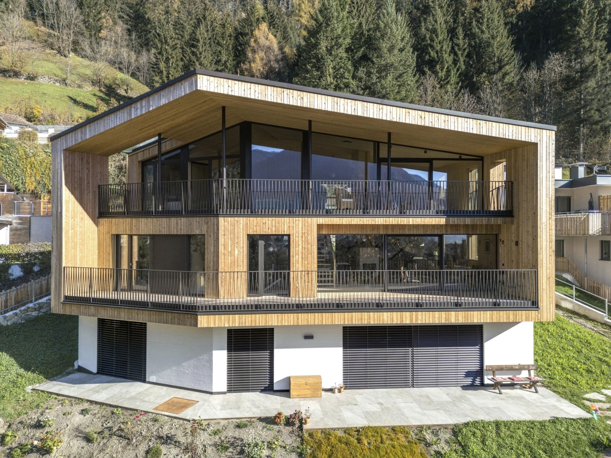 Una vista esterna di una moderna casa Holzius con facciata in legno e ampie vetrate. L'edificio presenta una forma geometrica distintiva, con un tetto spiovente e diversi balconi circondati da una ringhiera nera. In primo piano si vedono una zona pavimentata e un piccolo giardino con piante verdi. Gli alberi e le colline circostanti creano uno sfondo naturale, mentre il cielo รจ limpido e la scena risplende di una luce calda.
