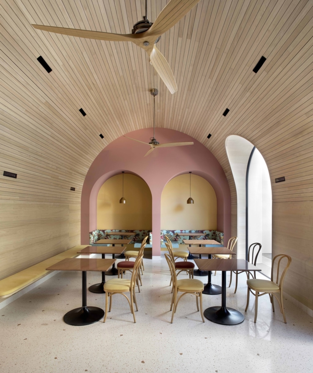 Moderna area caffetteria o salotto caratterizzata da un'architettura lineare e armoniosa e da caldi materiali naturali: diversi tavolini quadrati con basi rotonde nere e sedie in legno chiaro sono distribuiti in modo uniforme nella stanza. Sullo sfondo, due nicchie semicircolari incastonate in una parete color rosa, ciascuna dotata di panche imbottite e discrete lampade a sospensione. Il soffitto è interamente rivestito con sottili listelli di legno e si inarca dolcemente sopra la stanza, completato da due grandi ventilatori a soffitto con pale curve in legno. A sinistra, una panca continua si estende lungo la parete, mentre a destra un'alta finestra ad arco garantisce la luce naturale e la vista sull'esterno. Il pavimento chiaro e screziato, insieme alla combinazione di legno, morbide curve e tonalità calde, crea un'atmosfera tranquilla, accogliente e di grande pregio architettonico.