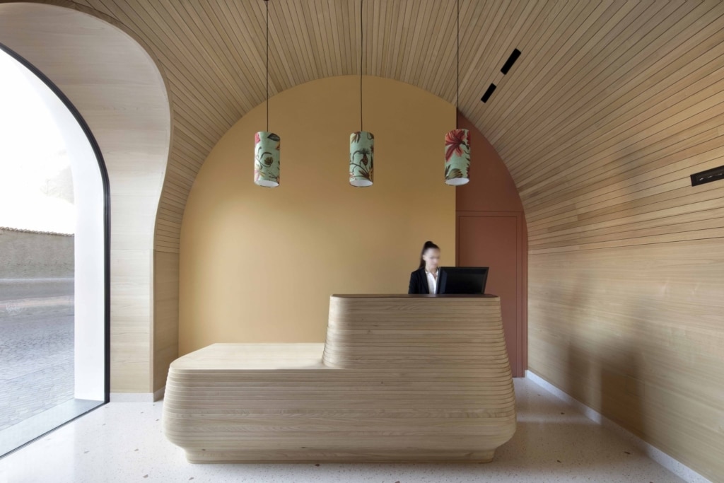 Vista dettagliata della zona reception di un interno dal design moderno, caratterizzato da un linguaggio formale chiaro e fluido: al centro spicca un bancone della reception dalle forme scultoree, realizzato in legno chiaro con una struttura a strati orizzontali. Dietro di esso si trova una parete in una calda tonalità ocra chiara, che sfuma in una forma del soffitto dalle curve morbide. Il soffitto è interamente rivestito con sottili listelli di legno e si estende con una curvatura uniforme sopra la stanza. Tre lampade a sospensione con paralumi cilindrici in tessuto a motivi floreali pendono simmetricamente sopra il bancone. A sinistra, una grande apertura in vetro a forma di arco garantisce la luce naturale e offre una vista sull'esterno. A destra, un rivestimento in legno continuo si estende lungo la parete. Dietro il bancone, una persona è in piedi davanti a uno schermo, a indicare la funzione di area reception. La combinazione di calde superfici in legno, morbide curve e design essenziale crea un'atmosfera tranquilla, raffinata e accogliente.