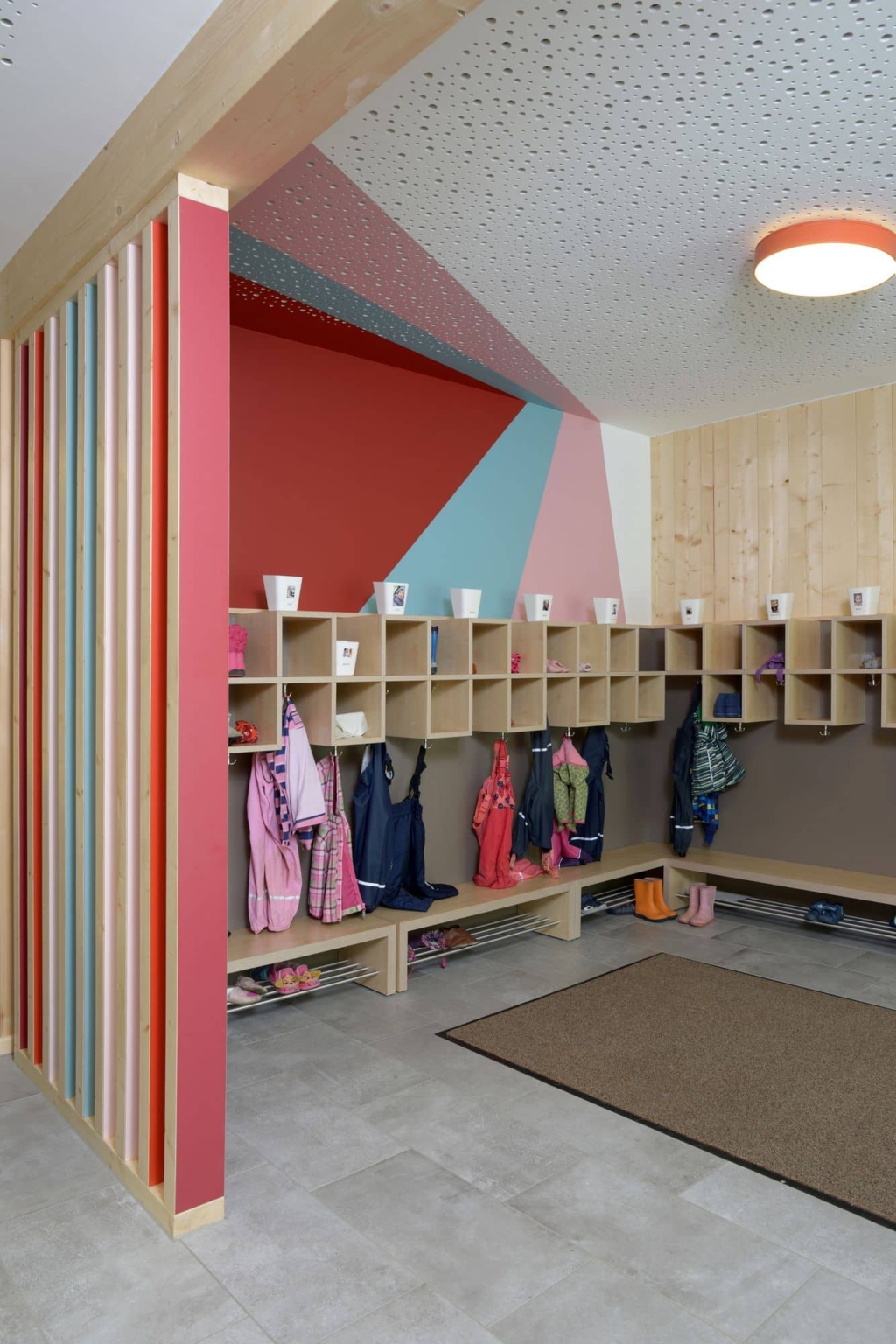 Un guardaroba dal design moderno e vivace, con rivestimenti in legno, ripiani aperti e ganci per i vestiti dei bambini, abbinato a pareti dai colori geometrici e a un'atmosfera accogliente e a misura di bambino.