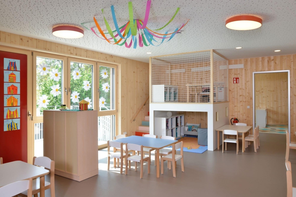 Una stanza per bambini moderna e luminosa, con rivestimenti in legno, ampie finestre, tavolini e sedie, decorazioni colorate e aree gioco e relax integrate, il tutto in un'atmosfera accogliente.