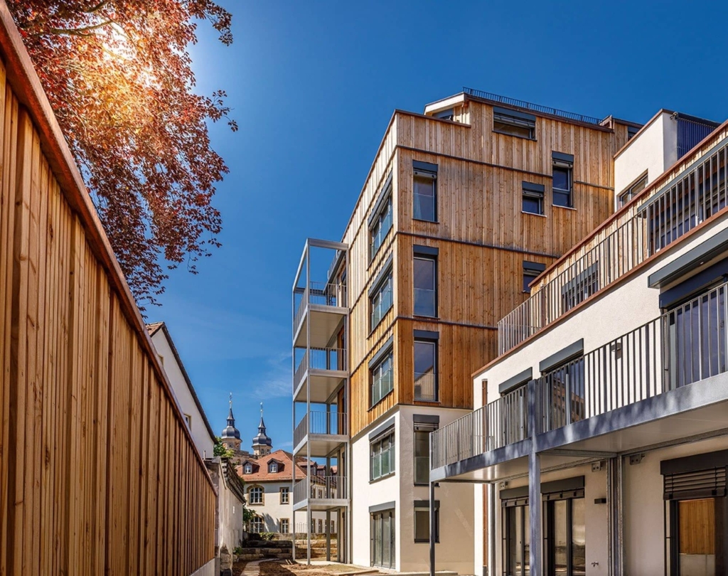 Un edificio moderno con una facciata in legno ed elementi bianchi sotto un cielo blu. A sinistra dell'immagine si vede un albero dalle foglie rosse. Sullo sfondo si intravede il campanile di una chiesa. Lo stile architettonico Holzius potrebbe riflettersi nella struttura in legno dell'edificio.