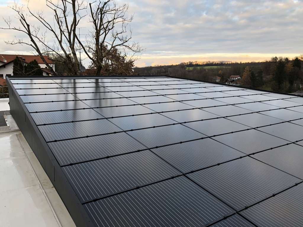 Una vista dal tetto di una casa moderna con un'ampia superficie di pannelli solari. I pannelli solari neri sono distribuiti uniformemente sul tetto e riflettono la luce del sole al tramonto. Sullo sfondo si intravedono alberi spogli, mentre il paesaggio digrada dolcemente. Il cielo è nuvoloso, con tonalità tenui che creano un'atmosfera tranquilla e serena.