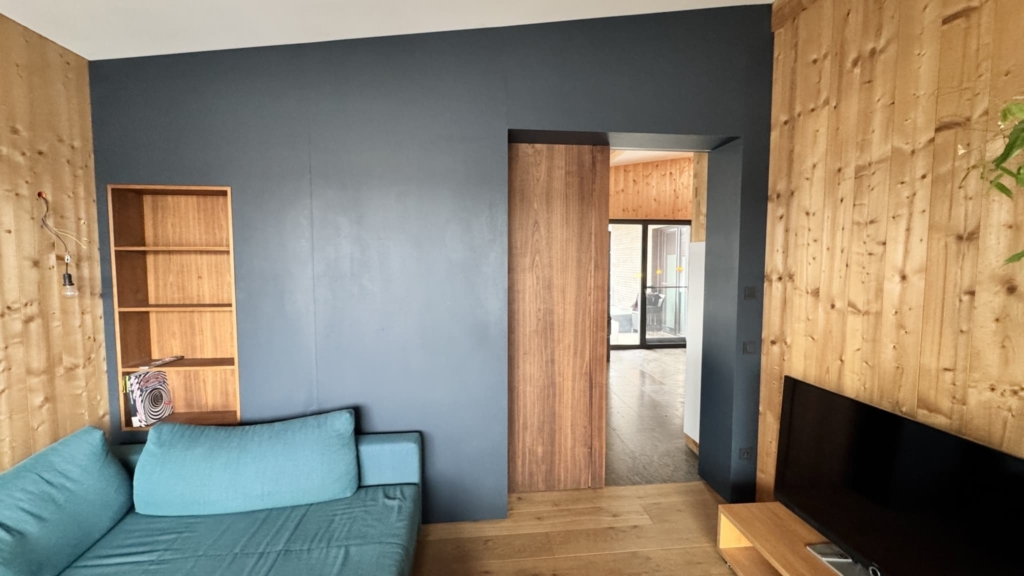 Una vista interna di un soggiorno con pareti in legno a vista. La parete di sinistra è rivestita con pannelli di legno chiaro, mentre quella di destra è dipinta in una tonalità di grigio intenso. In primo piano si trova un divano turchese e sulla parete è presente una libreria a scaffali aperti in legno. Sul lato destro è visibile una porta scorrevole in legno che conduce a un'altra stanza. Un televisore è appoggiato su un mobile in legno e il pavimento è costituito da assi di legno chiaro che creano un'atmosfera calda.