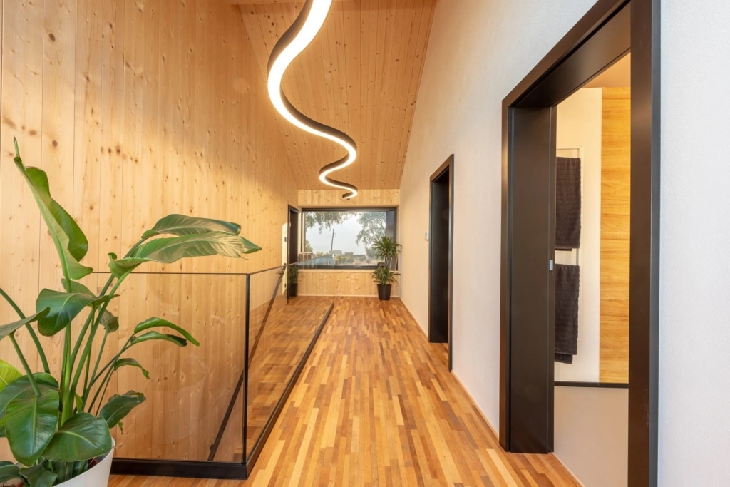 Una moderna casa modello con elementi in legno a vista su pareti e soffitto. Le pareti sono rivestite in legno chiaro, mentre il soffitto presenta un'elegante forma ondulata. Il pavimento รจ costituito da calde assi di legno. Una raffinata striscia luminosa ricurva pende dal soffitto e illumina la stanza. Sul lato sinistro si trova un vaso con una pianta verde, mentre sullo sfondo si aprono ampie finestre che offrono una vista sui dintorni.