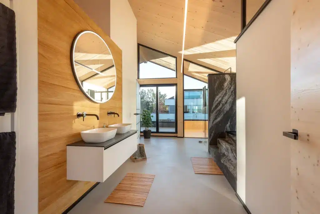 Un bagno moderno con un rivestimento in legno su una parete e un grande specchio rotondo sopra un lavabo sospeso con due vasche bianche. Le pareti sono chiare e il soffitto รจ alto e spiovente, con travi a vista. Una grande vetrata offre una vista sull'esterno, mentre il pavimento รจ realizzato in un materiale chiaro e liscio. Sul pavimento sono disposti due tappeti da bagno rotondi.