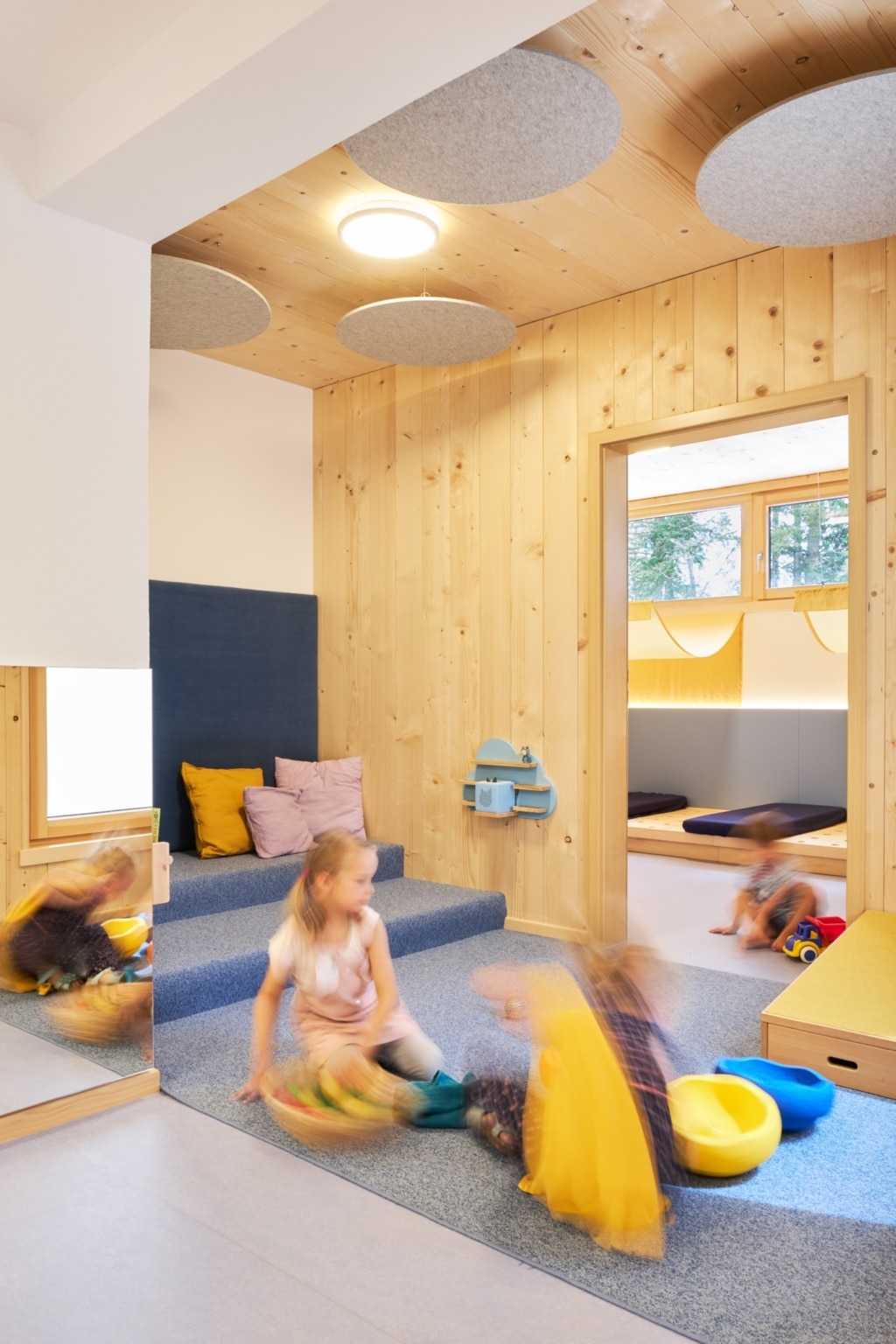Un interno luminoso e accogliente, con rivestimenti in legno a tutta superficie realizzati con elementi Holzius e un'atmosfera confortevole: una zona salotto imbottita con cuscini si apre su un'area giochi aperta, mentre elementi acustici rotondi sul soffitto garantiscono una piacevole acustica dell'ambiente. Attraverso un'ampia apertura si intravede un'altra sala relax arredata in modo morbido con zone relax ribassate; diversi bambini utilizzano lo spazio per giocare e muoversi.