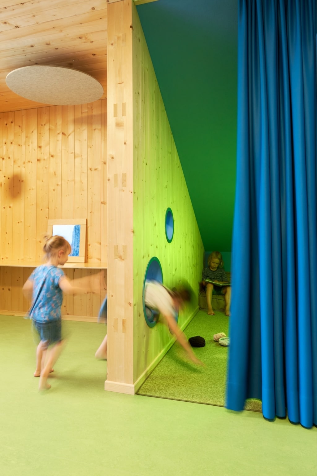 Un ambiente luminoso e ben strutturato, con rivestimenti in legno su tutta la superficie e materiali dalle tonalità calde: un’area giochi aperta, dotata di scaffali e ripiani integrati, si fonde con una zona colorata a forma di nicchia. L’angolo giochi, decorato in verde con aperture circolari nella parete e un morbido rivestimento del pavimento, offre al contempo un rifugio e uno spazio per giocare, mentre una tenda permette di isolare l’area in modo flessibile. Un bambino è seduto nella nicchia e guarda un libro, mentre un altro attraversa giocosamente la stanza.