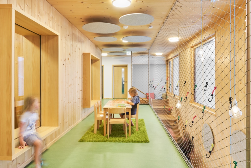 Un ambiente interno inondato di luce, con calde superfici in legno e un design essenziale: un ampio corridoio funge da area giochi e ricreativa, con nicchie di seduta integrate e una grande vetrata. Al centro, su un tappeto verde, c'è un tavolino dove un bambino sta giocando, mentre lungo il lato una rete da arrampicata tesa con elementi colorati offre ulteriore spazio per muoversi; pannelli acustici a soffitto garantiscono una piacevole acustica dell'ambiente.