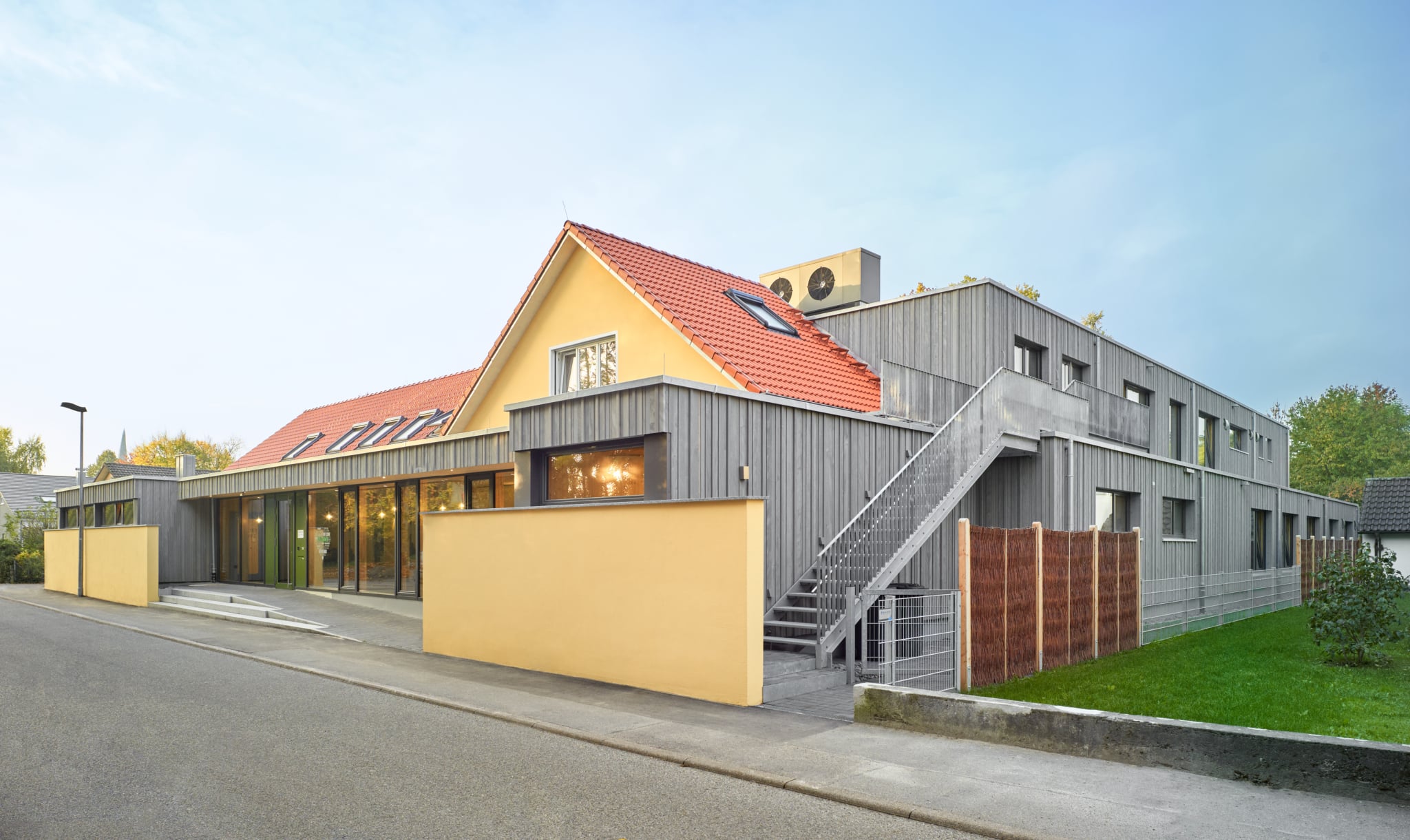 Un moderno progetto Holzius che vede un edificio esistente intonacato di giallo e con tetto a due falde rosso, integrato da una nuova costruzione in legno dalla struttura ben definita. L'ampliamento a due piani con facciata verticale in legno grigio presenta ampie superfici vetrate, una scala esterna e un linguaggio formale essenziale e contemporaneo. Sul lato strada, le facciate vetrate e l'area d'ingresso caratterizzano l'aspetto esterno, mentre sul lato si affaccia una piccola area giardino recintata.