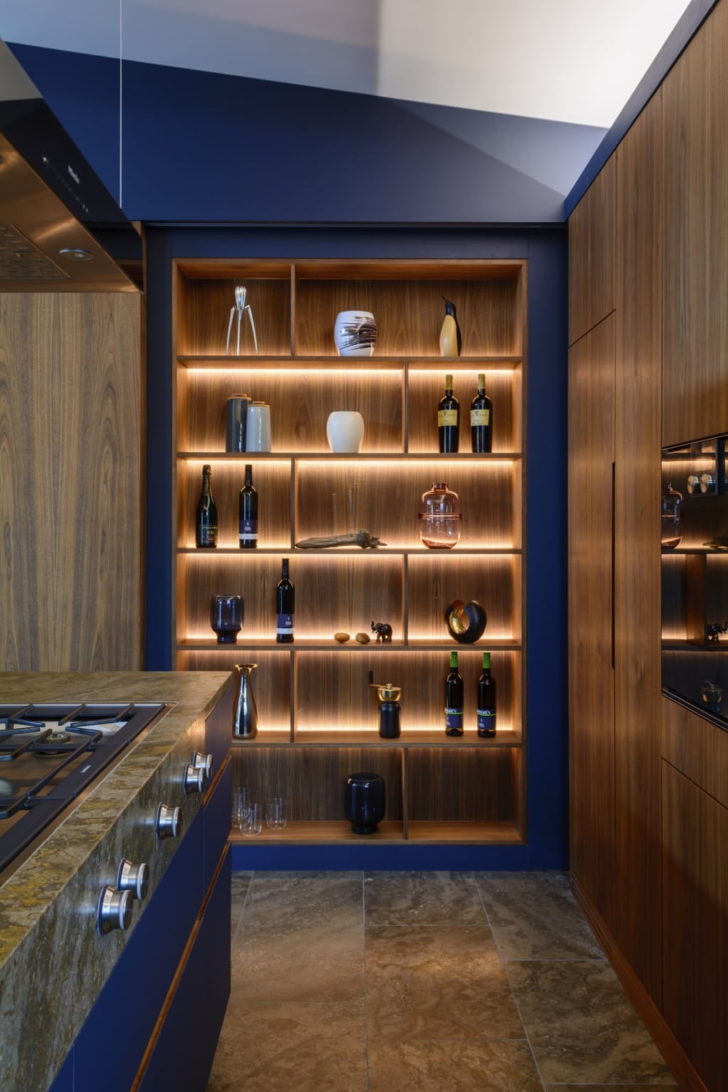 Una cucina moderna con una scaffalatura aperta in legno, illuminata da calde luci a LED. La scaffalatura è piena di bottiglie, vasi e oggetti decorativi. In primo piano si trova un piano cottura dotato di un elegante piano cottura e di un piano di lavoro in granito. Le pareti sono dipinte di un blu intenso, mentre il rivestimento in legno degli armadietti mostra una venatura naturale. Il pavimento è in piastrelle di pietra dai toni neutri, il che crea un'atmosfera elegante e accogliente.