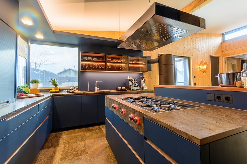 Una cucina moderna con una disposizione a L che combina elementi in legno e blu. L'isola centrale presenta un piano di lavoro robusto e un piano cottura integrato con manopole rosse. Sulla parete si trovano eleganti mobili con ripiani in vetro illuminati che mettono in risalto piatti e bicchieri. Ampie finestre lasciano entrare la luce del giorno e offrono una vista sui dintorni. Il pavimento è rivestito da piastrelle chiare che conferiscono alla stanza un'atmosfera elegante e accogliente.