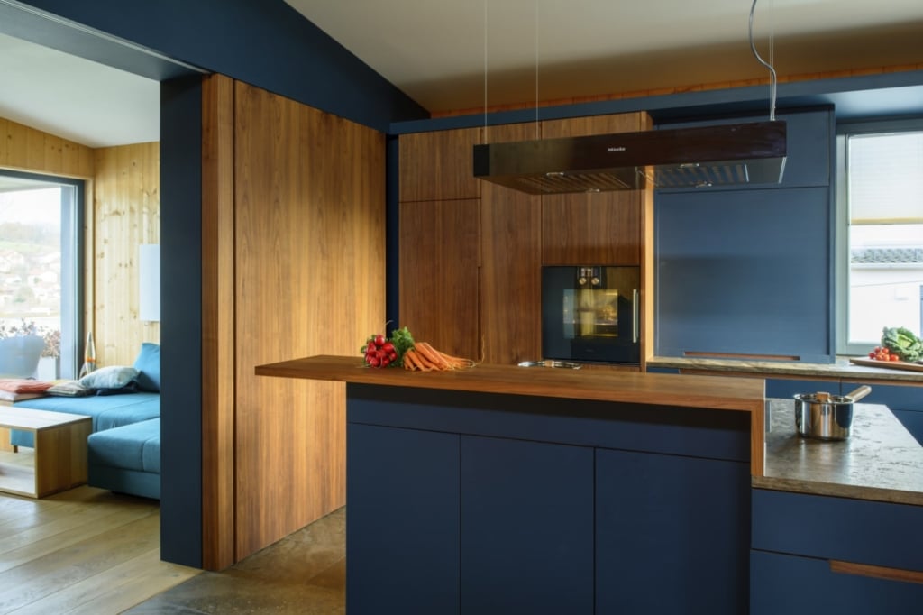 Una cucina moderna che combina superfici blu e in legno. In primo piano spicca un grande isola da cucina con piano di lavoro scuro e piano cottura integrato. Sulla parete è installata un'elegante cappa aspirante. Il rivestimento in legno sulla parete crea un caldo contrasto con i mobili blu. Sullo sfondo si intravede un accogliente salotto con un divano blu, che conferisce all'ambiente un aspetto invitante e moderno. La luce del giorno entra dalla finestra e illumina la stanza.