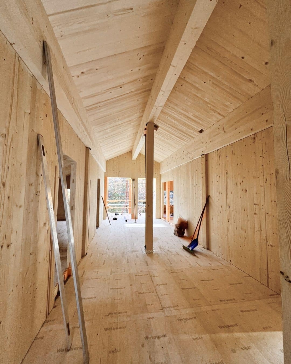 Interno della casa in legno massiccio in fase di costruzione, con travi a vista, pareti in legno massiccio e pavimento in legno; gli attrezzi da cantiere sono appoggiati alle pareti e la luce naturale filtra attraverso le ampie aperture in fondo alla stanza.
