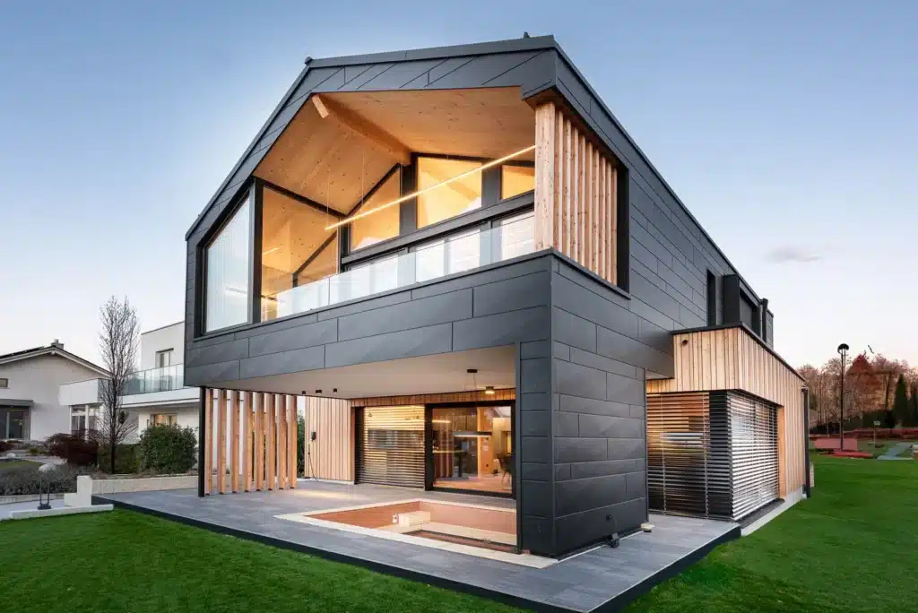 Una moderna casa modello caratterizzata da una combinazione di facciata nera e rivestimento in legno. L'edificio presenta una linea del tetto inclinata e distintiva e ampie finestre che lasciano entrare molta luce naturale. La facciata anteriore mostra una zona coperta con doghe in legno e un ingresso aperto con terrazza. Il prato davanti alla casa รจ ben curato e sullo sfondo si intravedono altri edifici residenziali.