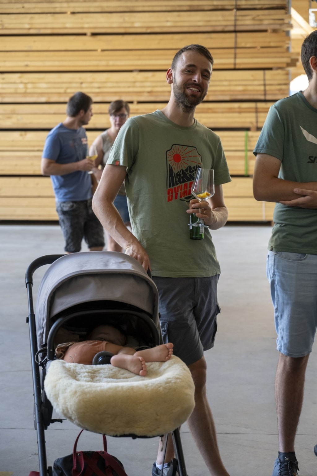 Mitarbeiter von holzius mit einem Kinderwagen, der ein leeres Aperitif-Glas in der Hand hält. Im Hintergrund sieht man ein paar Personen vorbei laufen sowie Holzstapel.