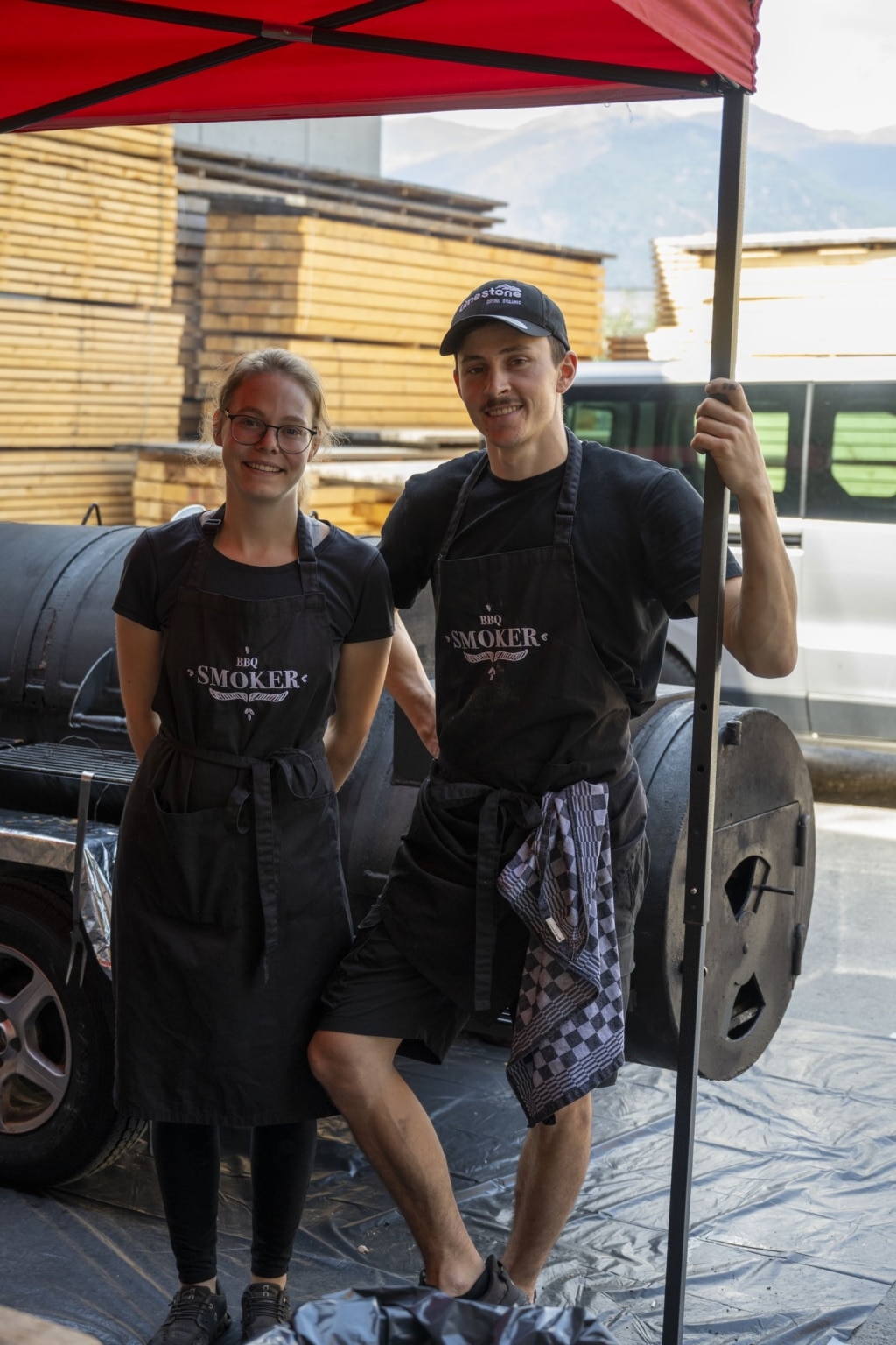 Grillmeister der Feier. Links steht eine junge Dame, die eine Brille trägt, rechts ein junger Herr. Beide tragen schwarz, sowie eine schwarze Schürze mit der Aufschrift BBQ Smoker.