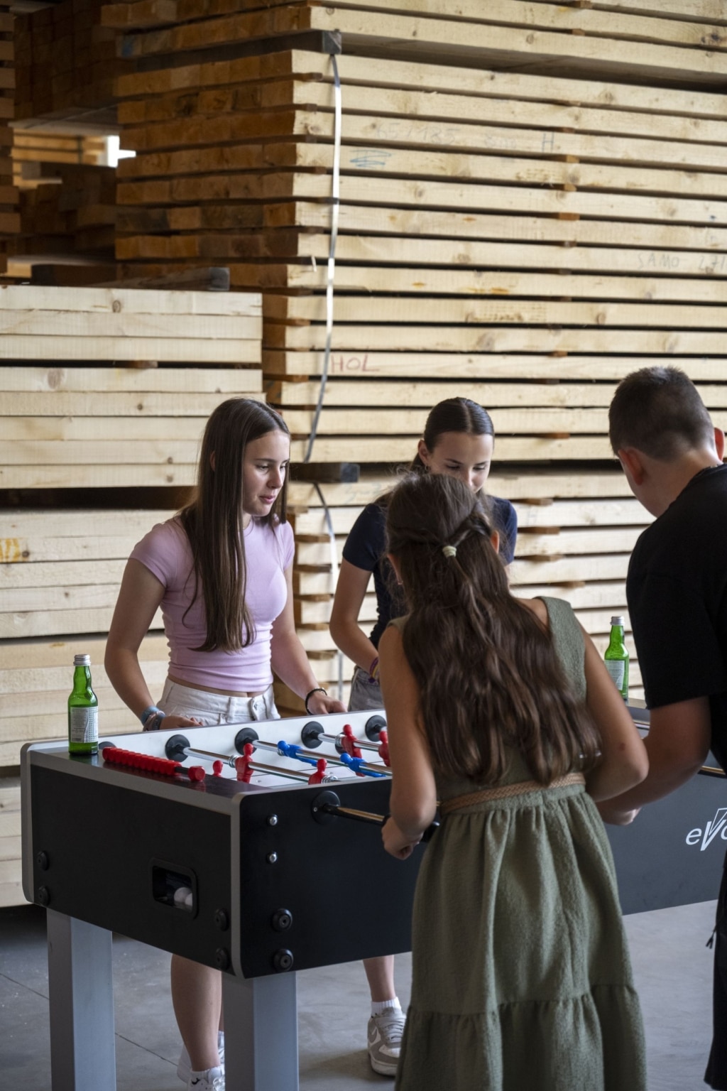Jungendliche die zusammen Tischfußball spielen. Im Hintergrund sieht man Holzstapel.