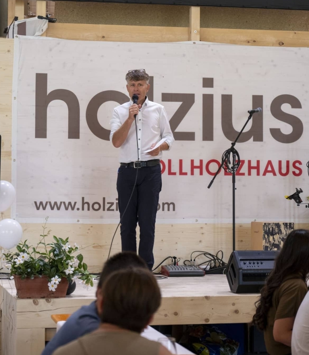 Bild von Herbert Niederfriniger, der auf der Bühne vor dem holzius Vollholzhaus Logo steht und eine Rede hält. Er hat ein weißes Hemd an und hält ein Mikrofon in der Rechten Hand und einen Zettel in der linken.