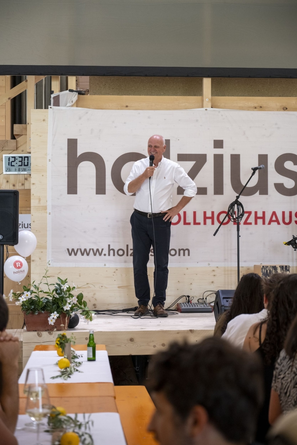Peter Rubner steht auf der Bühne und hält eine Rede. Er lächelt und trägt ein weißes Hemd. In seiner rechten Hand hält er ein Mikrofon und im Hintergrund sieht man das holzius Vollholzhaus Logo sowie www.holzius.com auf einer Wand. Die Bühne ist aus Holz.