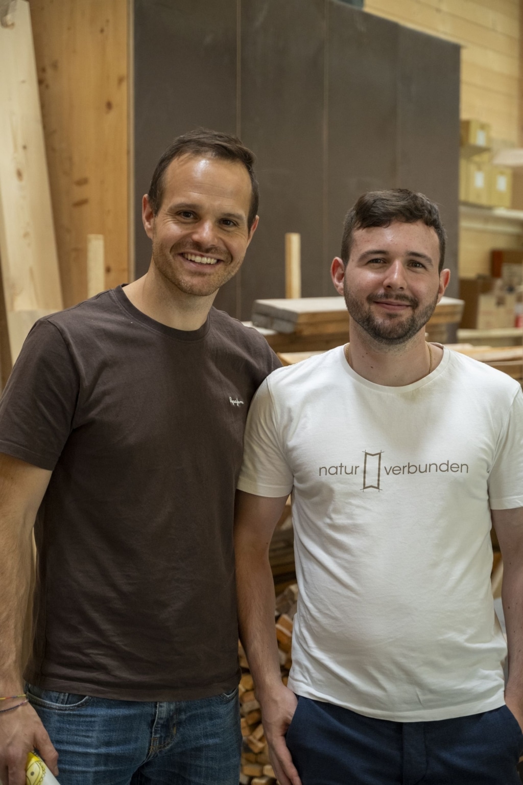 Zwei Mitarbeiter der holzius GmbH, die in die Kamera lächeln. Die linke Person trägt ein braunes Shirt und die rechte Person ein weißes Shirt mit der Aufschrift "naturverbunden" sowie der holzius-Gratleiste. Im Hintergrund sieht man viel Holz.