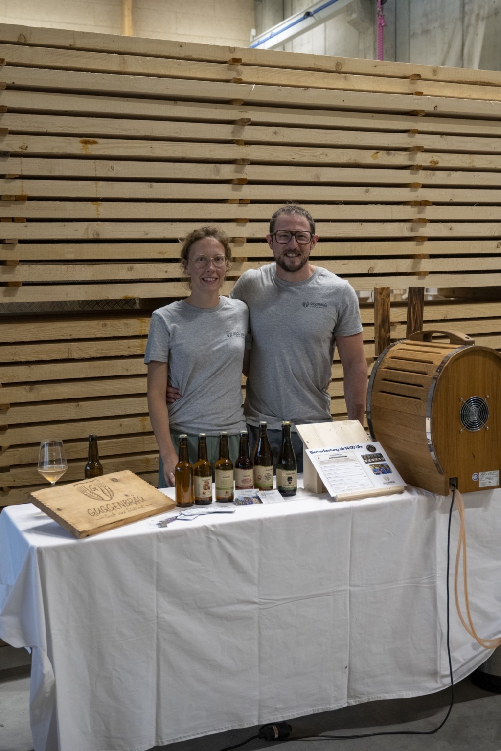 Inhaber der Brennerei Guggenbräu vor ihrem Ständchen mit den verschiedenen Biersorten. Im Hintergrund sieht man einen Holzstapel.