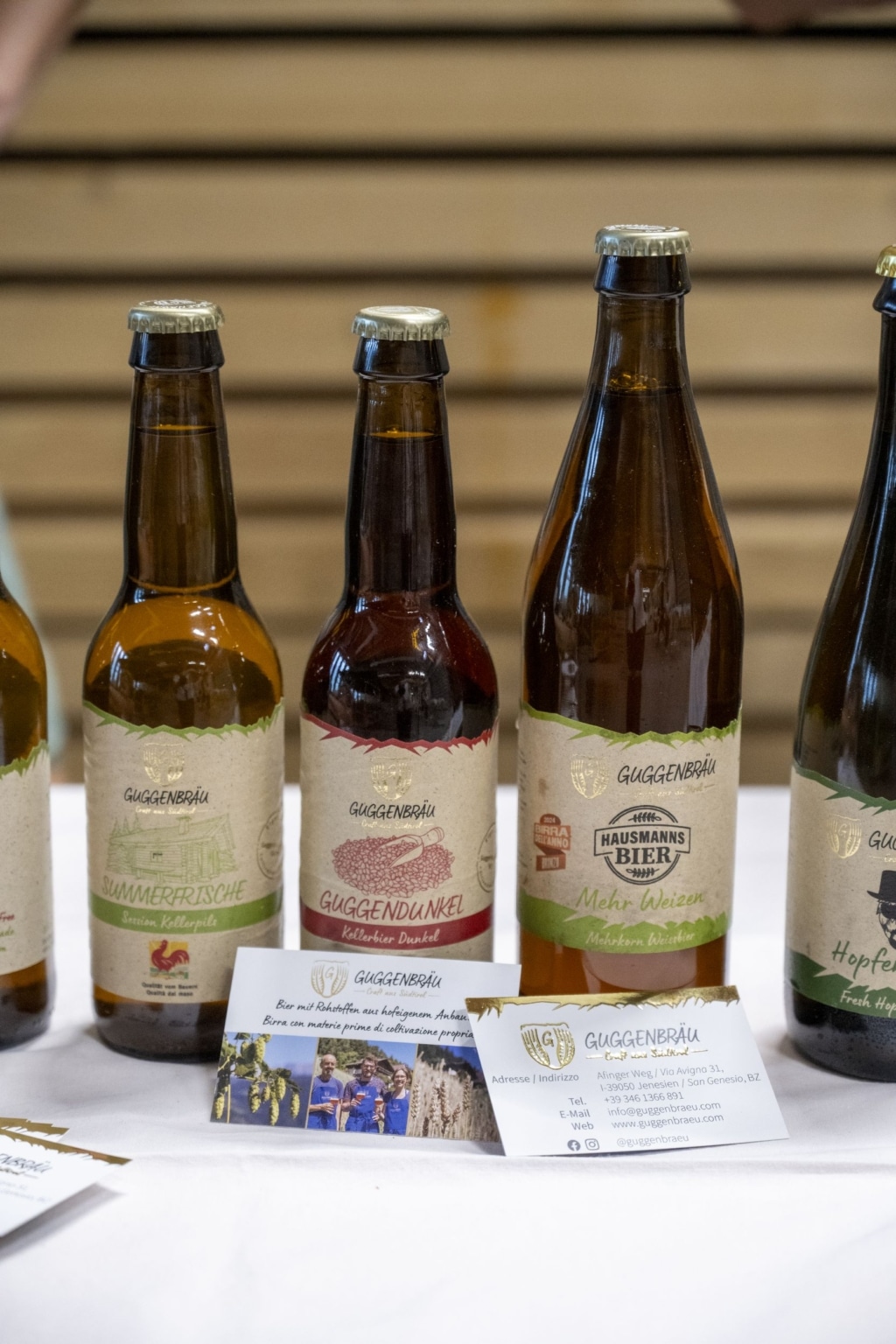 Flaschen mit verschiedene Biersorten von Guggenbräu, die auf einem Tisch mit weißer Tischdecke stehen. Davor liegen Kärtchen von Guggenbräu.
